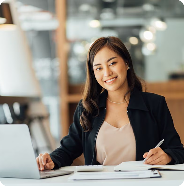 Smiling woman working at a desk with a laptop, notebook, and pen in a modern office setting.