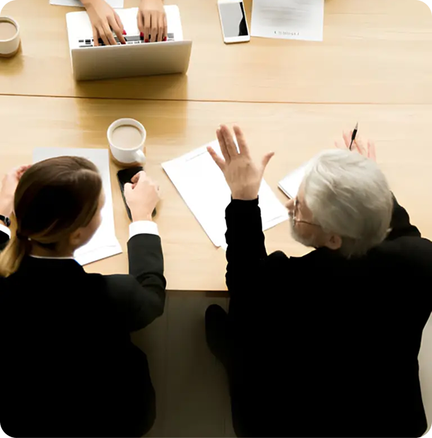 Overhead view of three professionals at a wooden table, one typing on a laptop, another with a coffee mug, and the third gesturing while holding a pen.