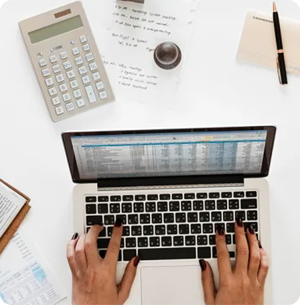 Overhead view of hands typing on a laptop keyboard surrounded by a calculator, papers, a stamp, and a pen on a white desk.