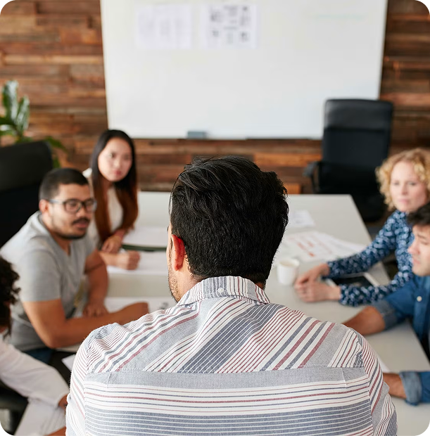 Group of five people having a meeting around a table in an office with a whiteboard in the background.