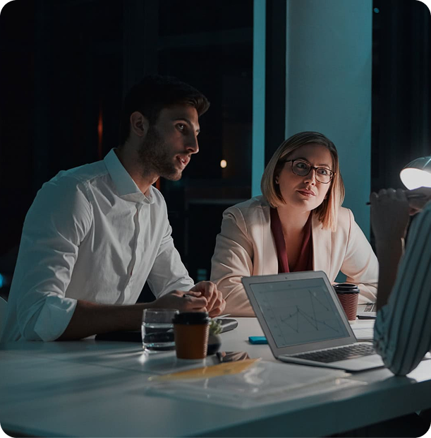 Three colleagues having a discussion at a desk with a laptop showing a graph during a nighttime office meeting.