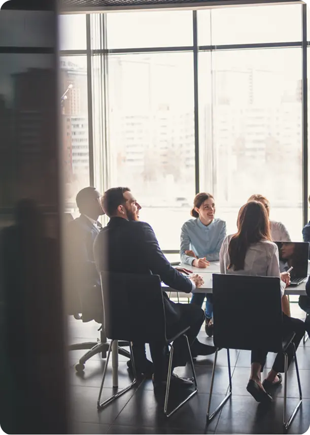 Business professionals sitting around a conference table engaged in a meeting near large windows with a city view.