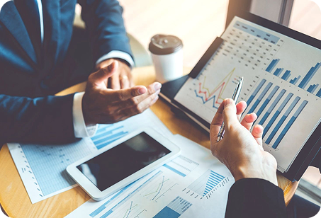 Two people reviewing financial charts and graphs on a tablet and printed documents at a wooden desk.