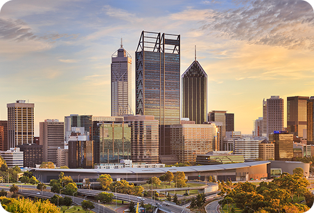 City skyline with modern skyscrapers and a sunset sky, featuring green trees and roads in the foreground.