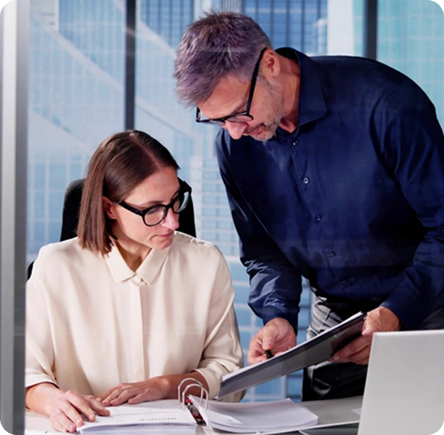A man and a woman wearing glasses review documents together in a modern office with large windows.