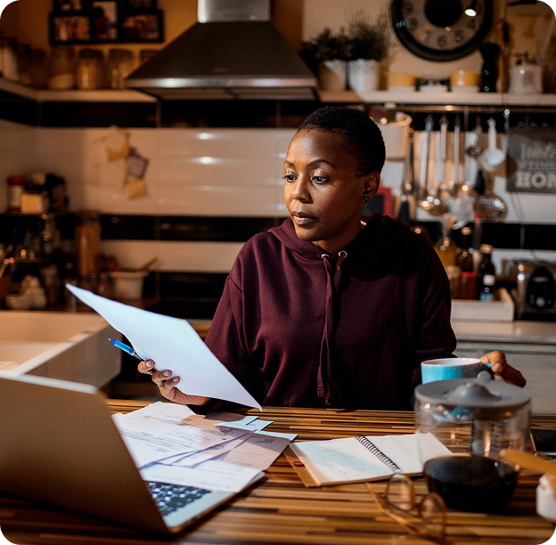 Woman in a maroon hoodie sitting at a kitchen table reviewing documents with a laptop and notebook nearby.