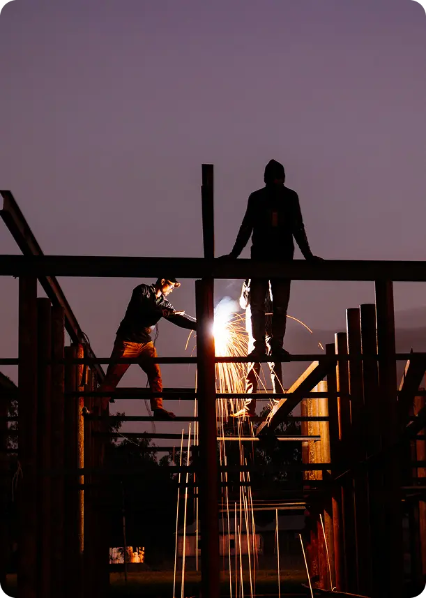 Two silhouetted workers performing welding at a construction site during dusk with bright sparks flying.