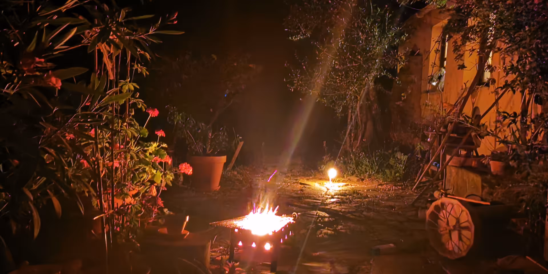 Frau sitzt bei Nacht mit ausgestreckten Beinen neben einem kleinen Grillfeuer in einem Garten mit Pflanzen und warmem Licht.