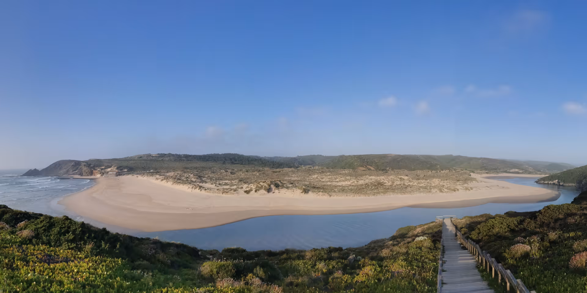 Strand mit goldenem Sand, ruhigem Meer und bewachsenen Klippen unter blauem Himmel mit Wolken.