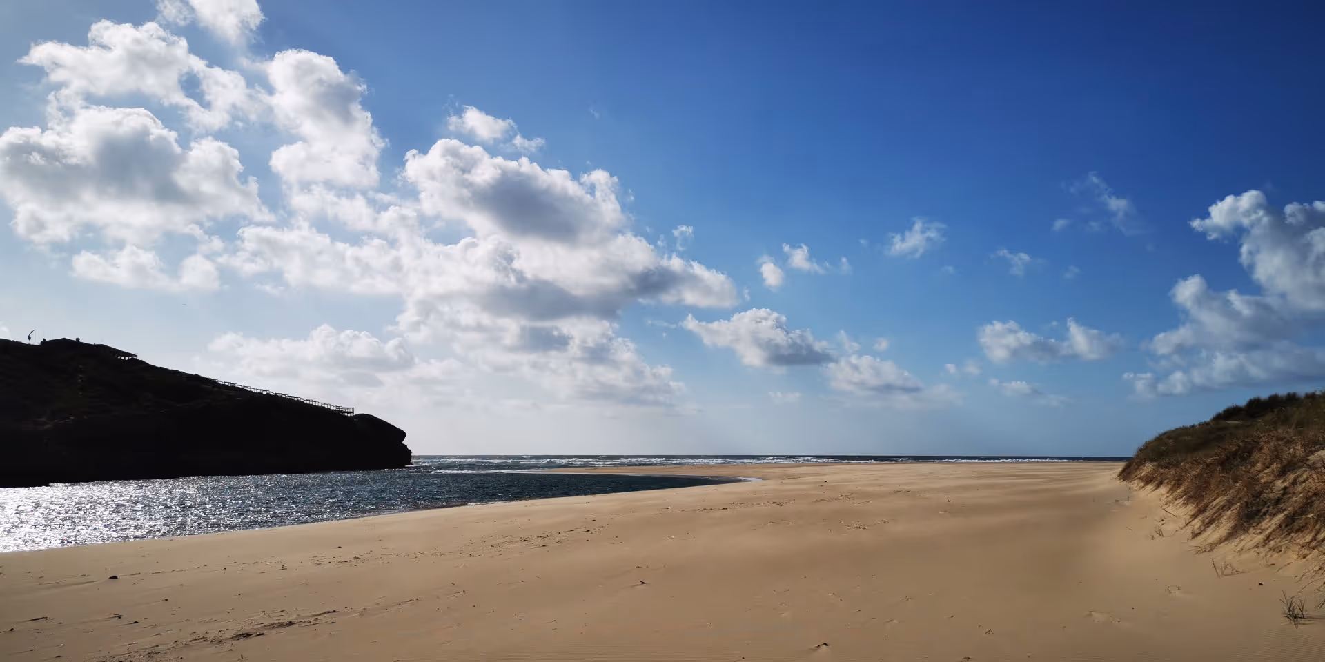 Luftaufnahme einer Küstenlinie mit blauem Meer, weißen Wellen und sandigem Strand vor bewachsenen Hügeln.