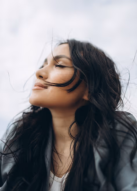 Woman with long dark hair and closed eyes enjoying the breeze against a cloudy sky.
