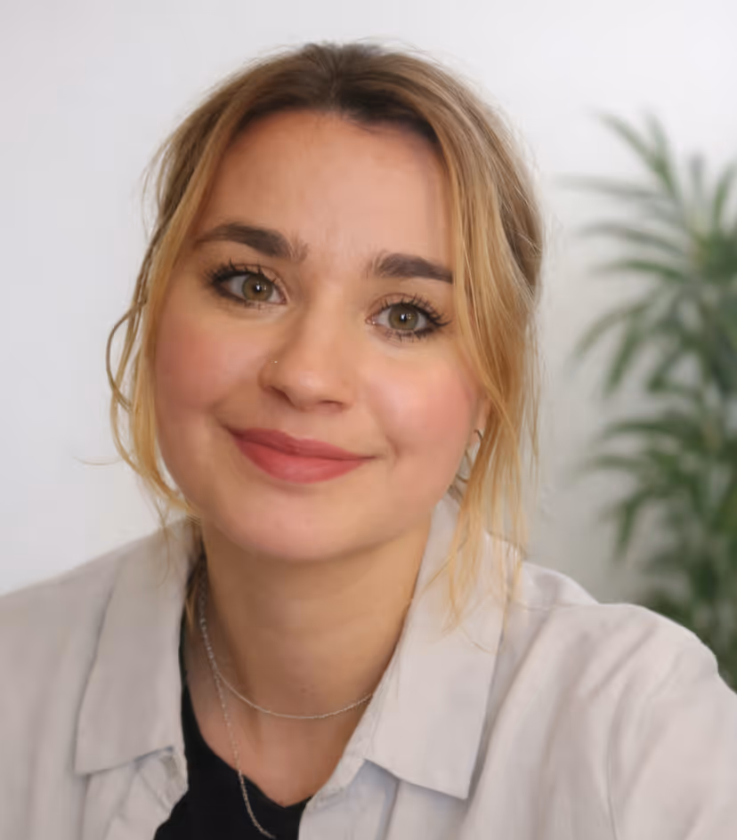 Smiling woman with blonde hair, wearing a light-colored shirt and black top, with a blurred green plant in the background.