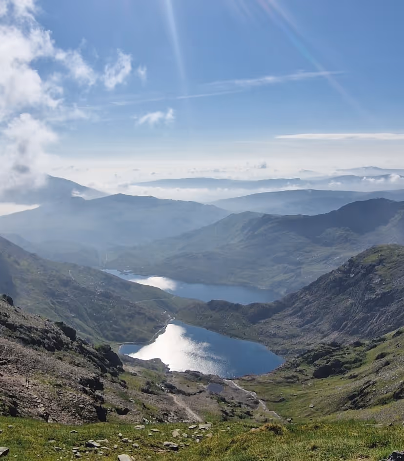 Mountain landscape with green slopes, rocky terrain, and two reflective lakes under a blue sky with scattered clouds.
