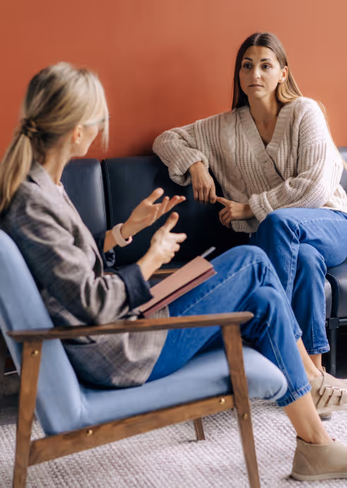 Two women sitting on a couch and chair in a conversation, one gesturing with hands while the other listens attentively.
