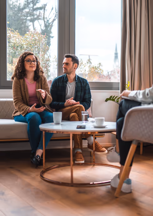 A couple sitting on a couch having a conversation with a therapist in a bright room.