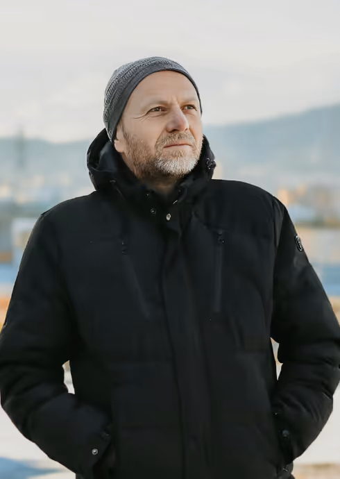 Middle-aged man with beard wearing a gray knit hat and black winter jacket, looking thoughtfully into the distance outdoors.