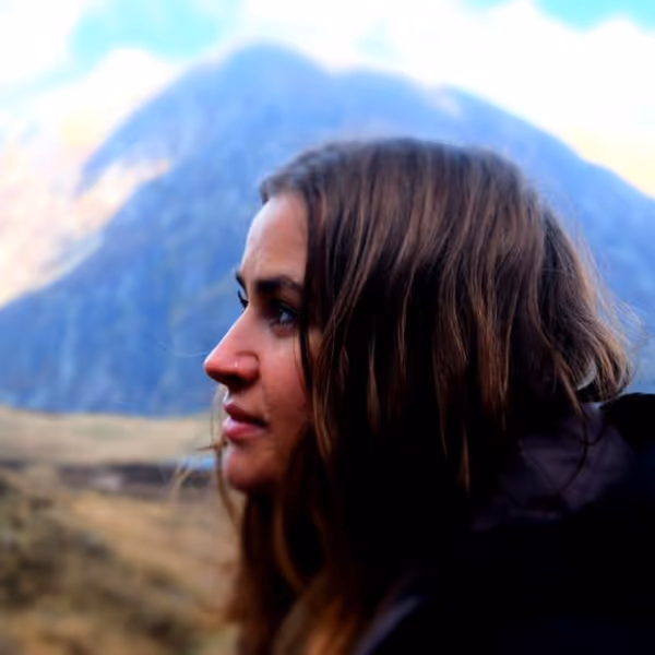 Side profile of a woman with long brown hair outdoors with a mountain and grassy landscape in the background.