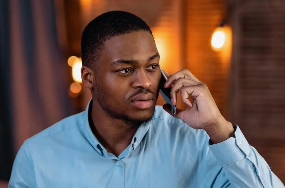 Young man in a light blue button-up shirt talking on a smartphone with a thoughtful expression indoors.