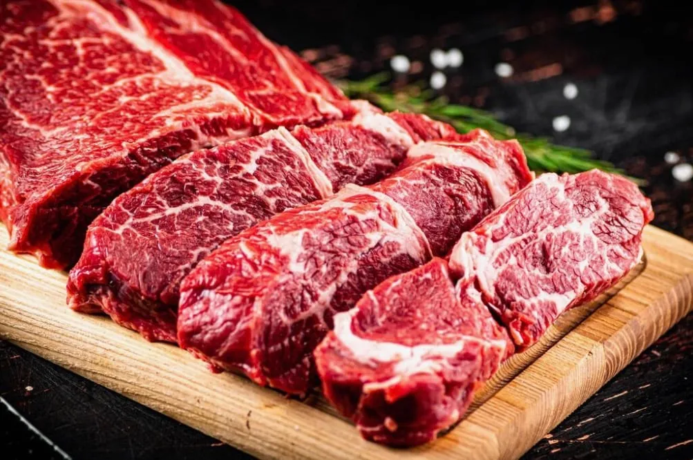 Fresh raw marbled beef steaks arranged on a wooden cutting board with a sprig of rosemary and coarse salt in the background.