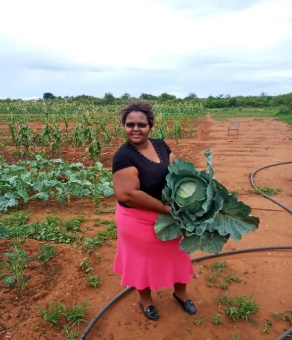 Woman standing in a field holding a large green cabbage, with crops growing in the background under a cloudy sky.