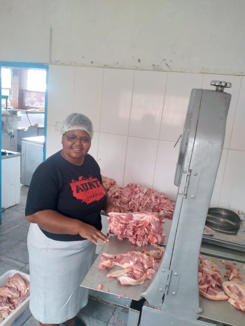 A woman wearing a hairnet, glasses, and a black shirt with 'AUNT Sakhe' printed on it, standing next to a large meat cutting machine with chunks of raw meat on the table.