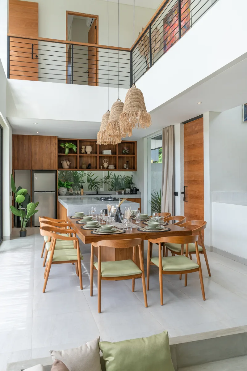 Modern dining room with large wooden table, eight chairs with green cushions, open kitchen in the background and wicker pendants in a Bali villa