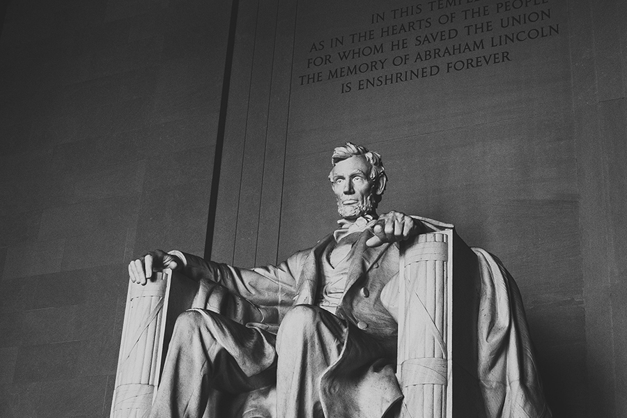 Black and white photo of the Abraham Lincoln statue seated inside the Lincoln Memorial with an inscription on the wall behind.