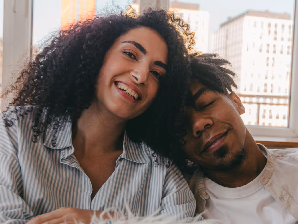 Smiling woman with curly hair leaning on a man's shoulder as he rests with eyes closed.