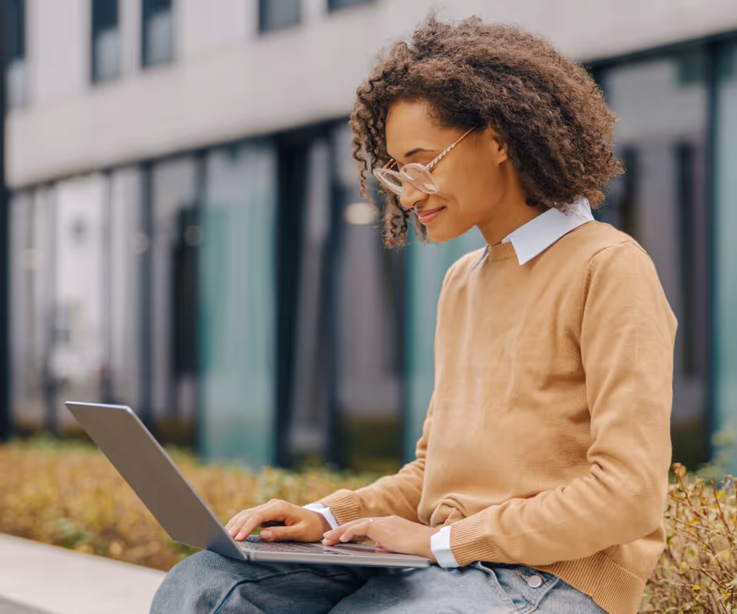 Young woman with curly hair and glasses typing on a laptop while sitting outdoors near a building.