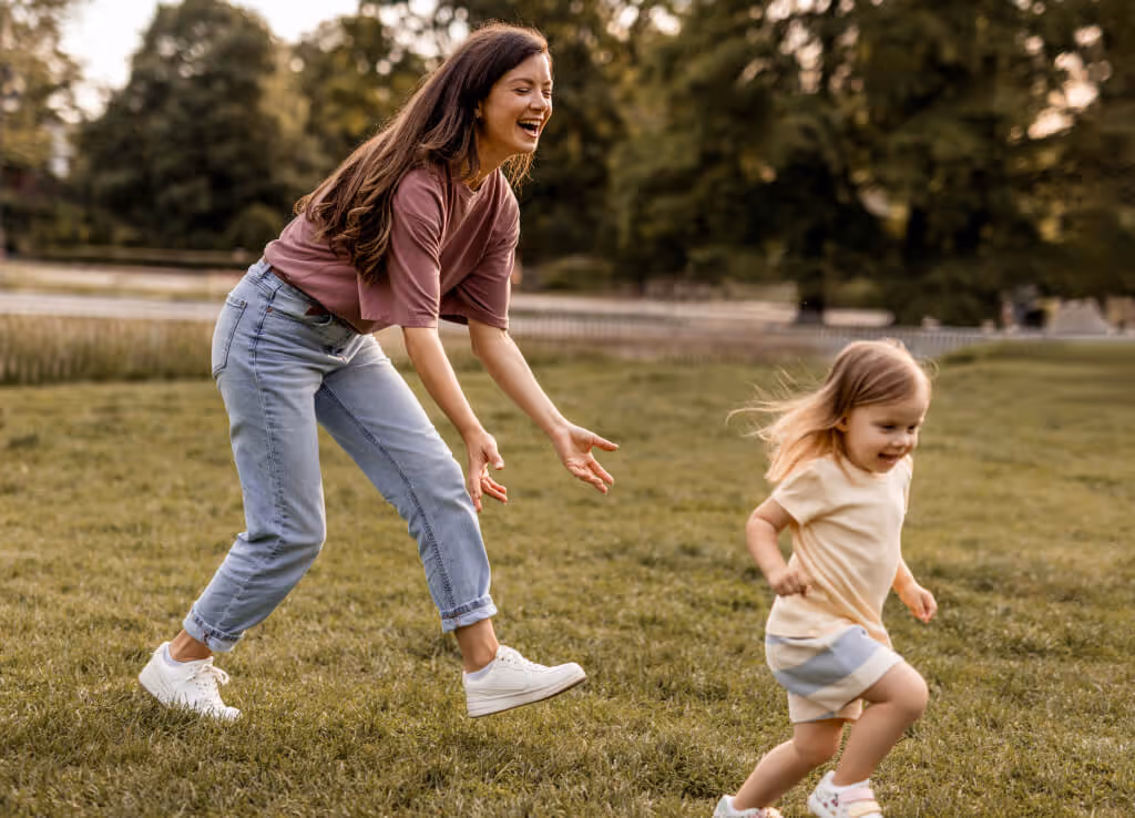 Smiling woman in pink shirt and jeans playing and chasing a young girl running on grass in a park.