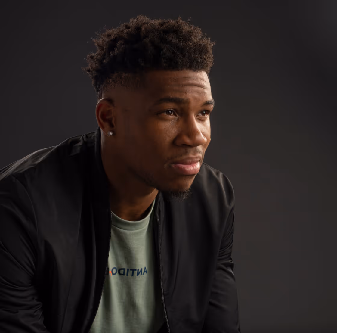 Young man with short curly hair and a stud earring, wearing a black jacket and green shirt, looking thoughtfully to the side against a dark background.