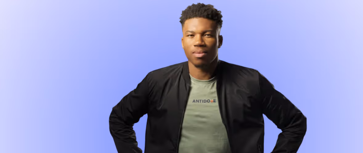 Young man with short curly hair wearing a black jacket over a green T-shirt standing with hands on hips against a purple background.