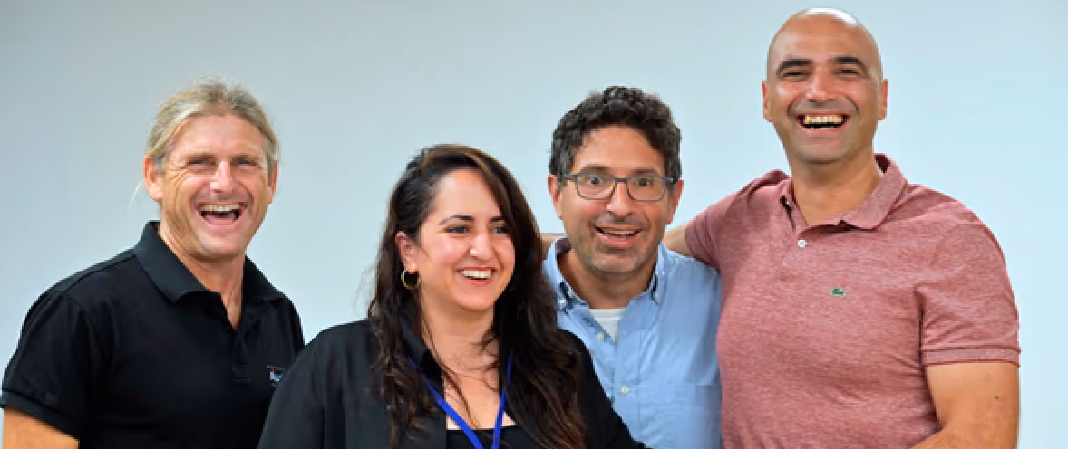 Group of four diverse smiling adults standing close together in front of a plain wall.