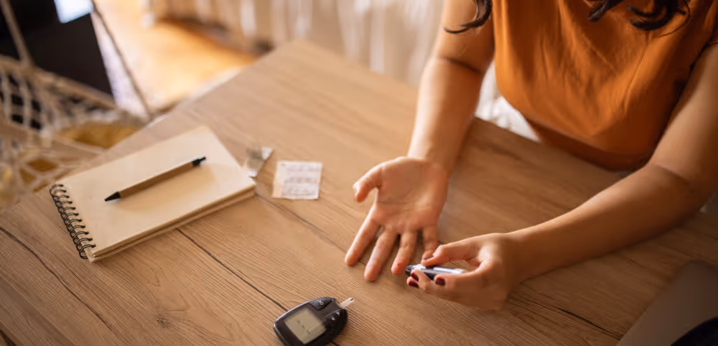 Person using a lancet device to test blood sugar on their finger with a glucometer, notebook, and test strips on a wooden table.