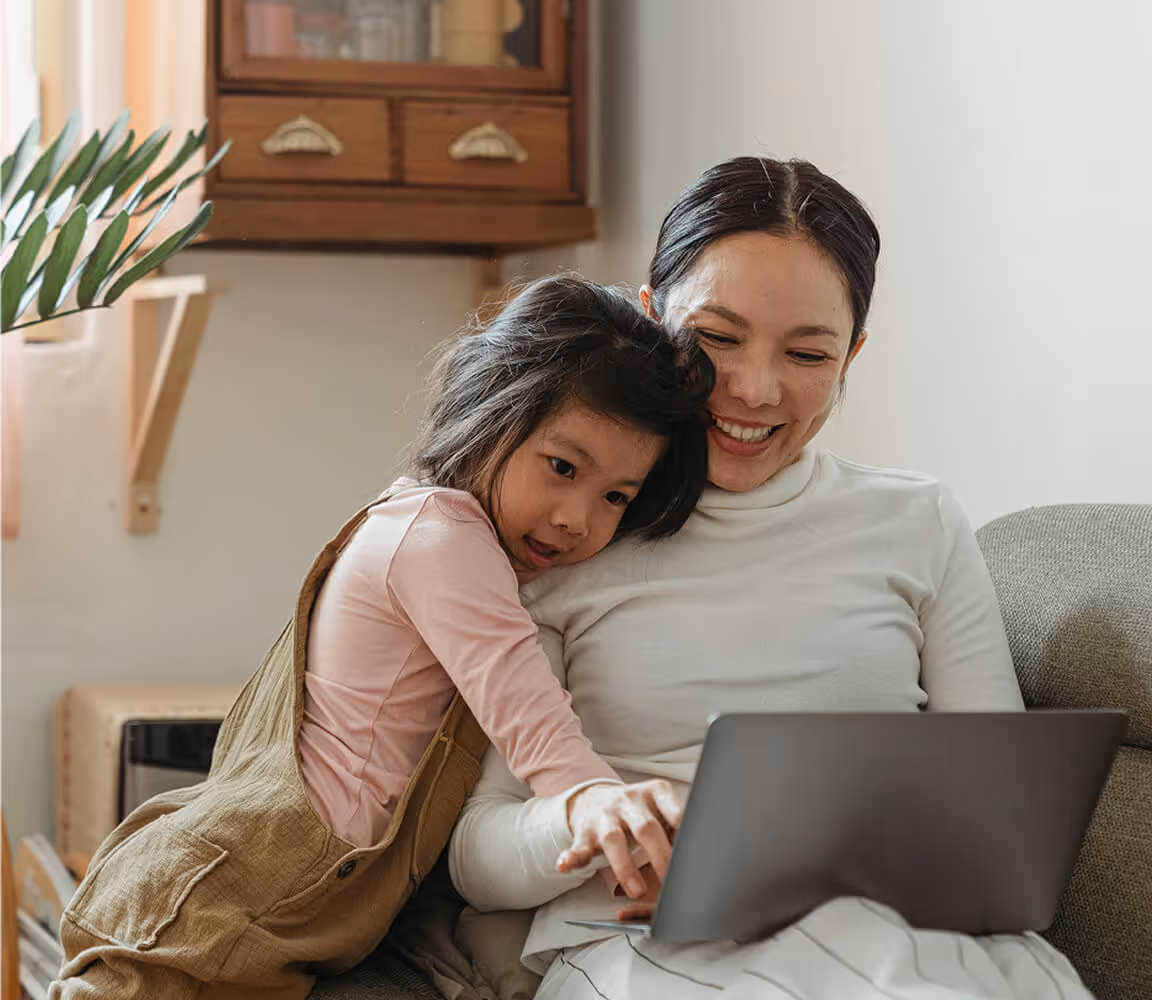 Mother and daughter smiling and using a laptop together on a couch in a cozy living room.
