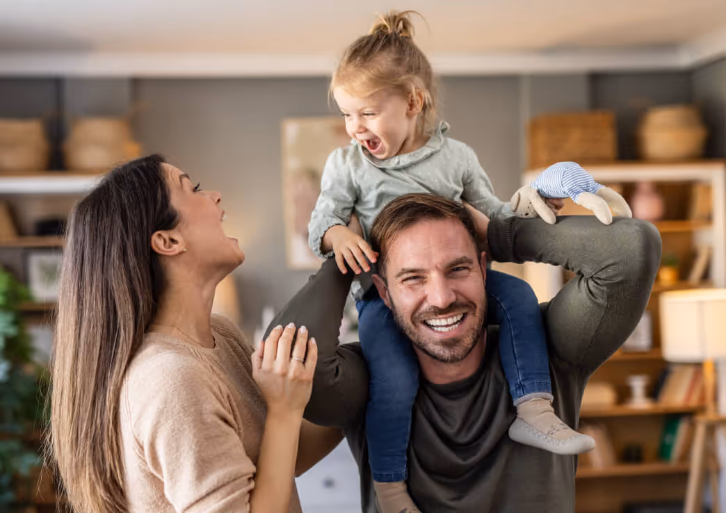 Smiling man carrying a laughing toddler on his shoulders while a woman joyfully interacts with them in a cozy living room.