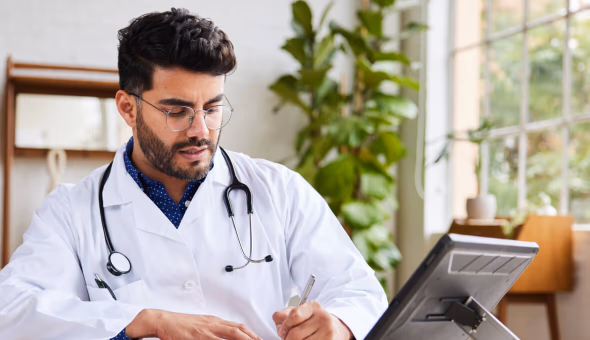 Male doctor wearing glasses and a white coat with a stethoscope around his neck writing notes at a desk with a tablet and green plants in the background.