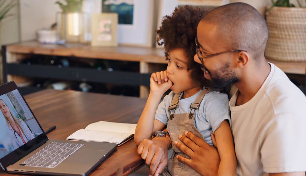 Father and child having a video call with a female doctor on a laptop at a home desk.