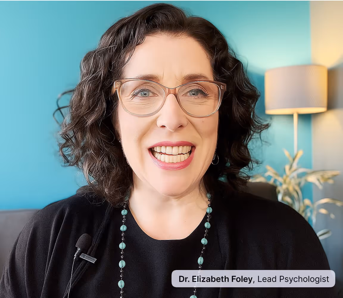 Smiling woman with curly dark hair and glasses wearing a black top and turquoise beaded necklace, with a teal wall and lamp in the background.
