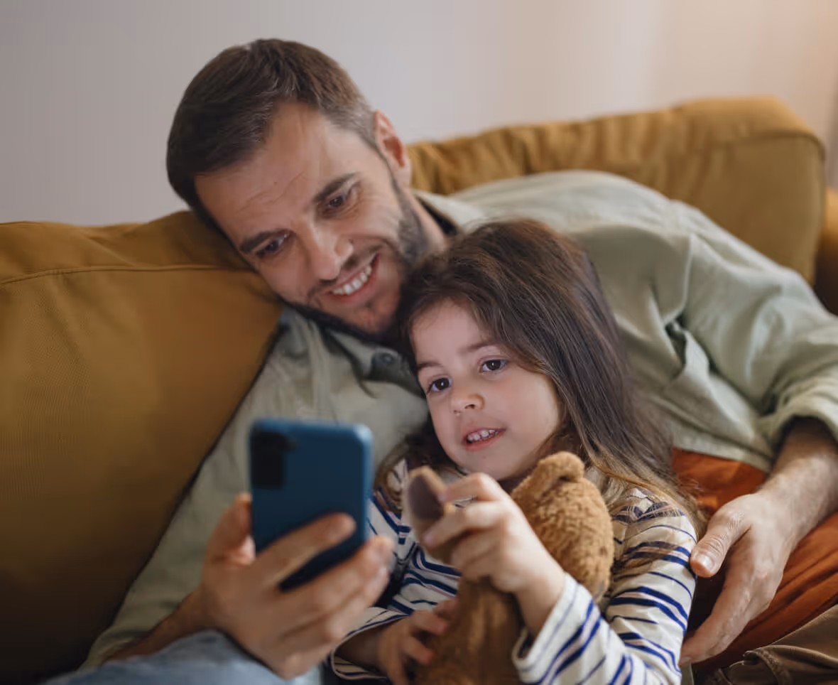 Smiling man and young girl sitting on a couch looking at a smartphone while the girl holds a teddy bear.