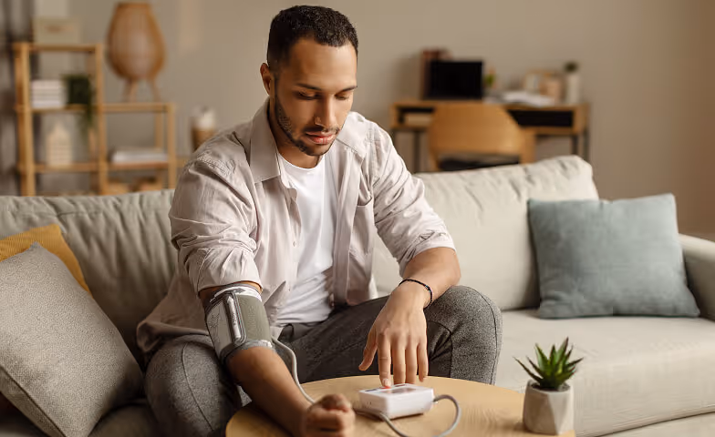 Young man sitting on a couch measuring his blood pressure using an arm cuff and a digital monitor on a table.