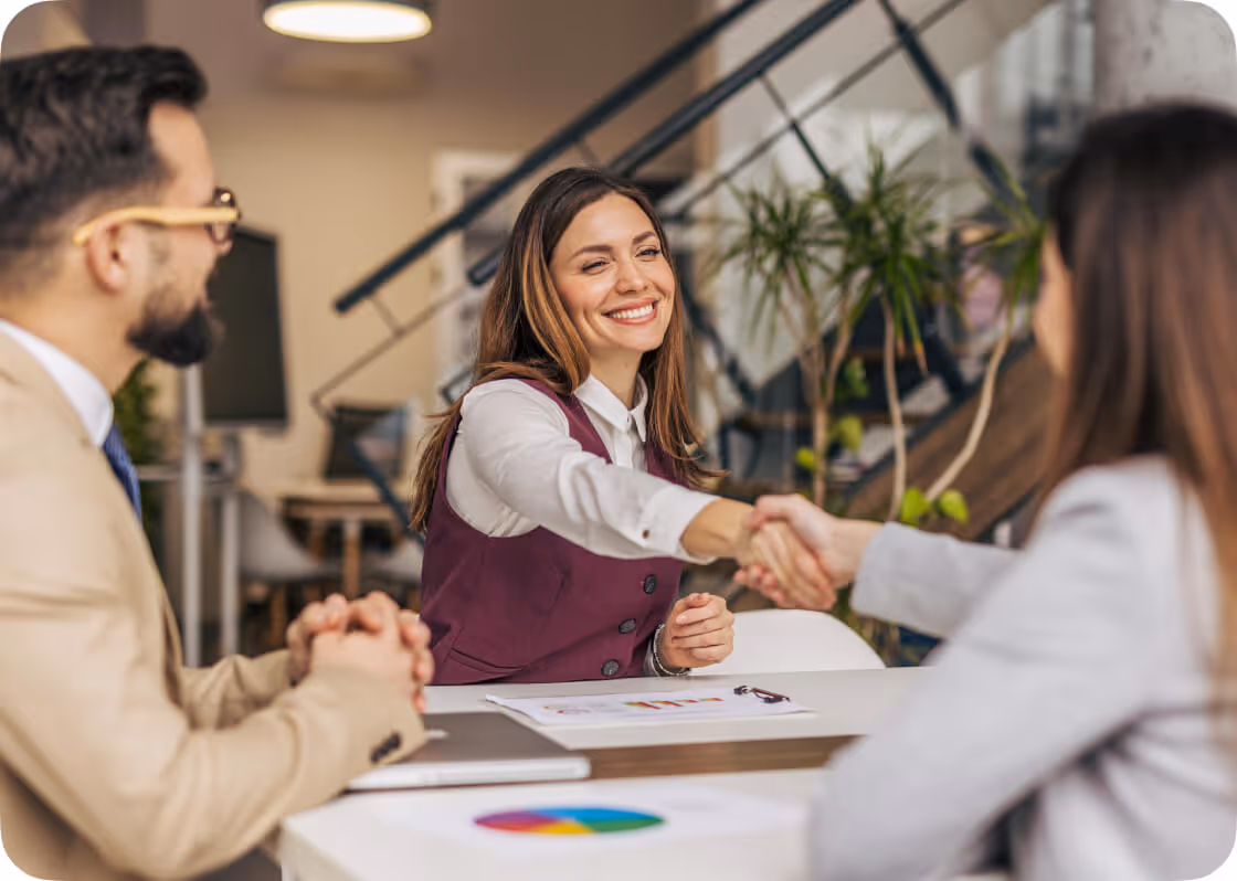 A smiling woman shaking hands with another person across a desk in an office meeting, while a man watches.
