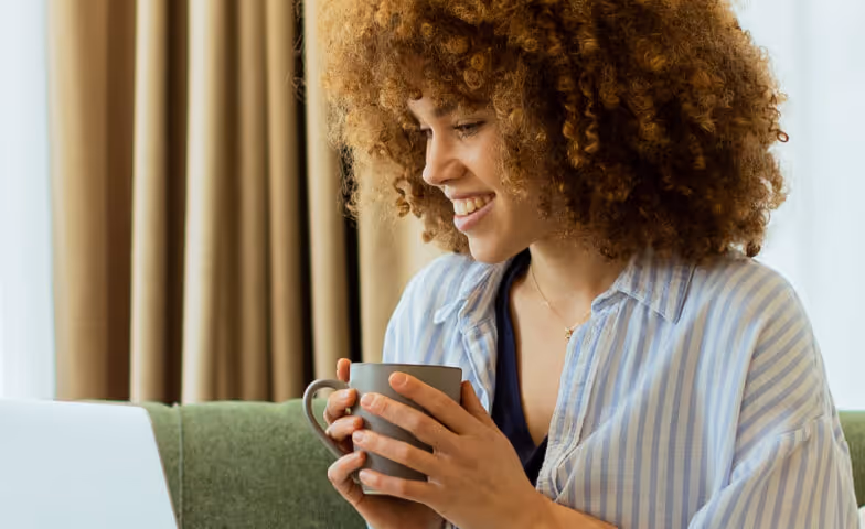 Smiling woman with curly hair holding a mug and looking at a laptop screen.
