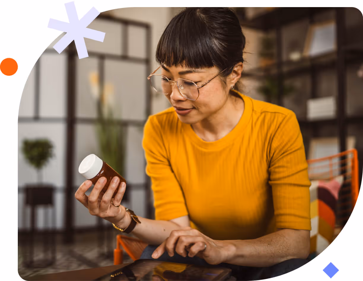 Woman in a yellow top holding a pill bottle and looking at a tablet indoors.