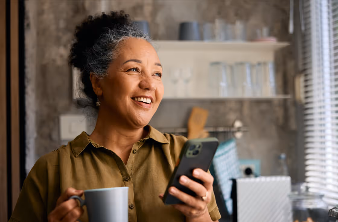Smiling woman holding a smartphone in one hand and a mug in the other in a kitchen setting.