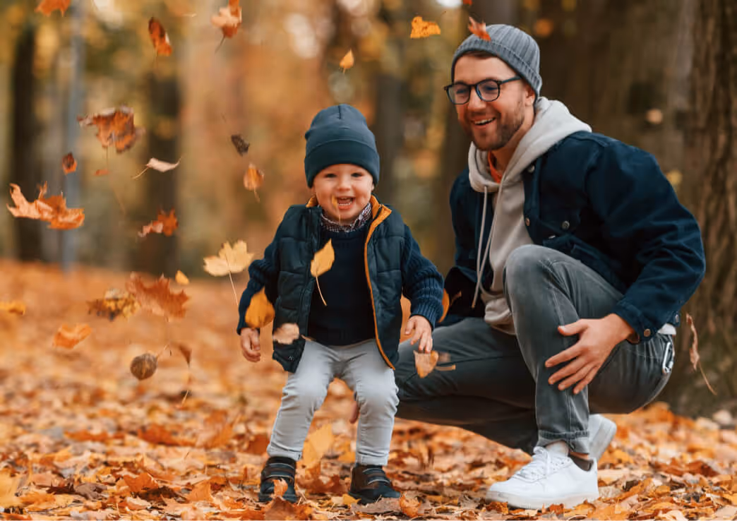 Smiling young child and man playing among falling autumn leaves in a forest.