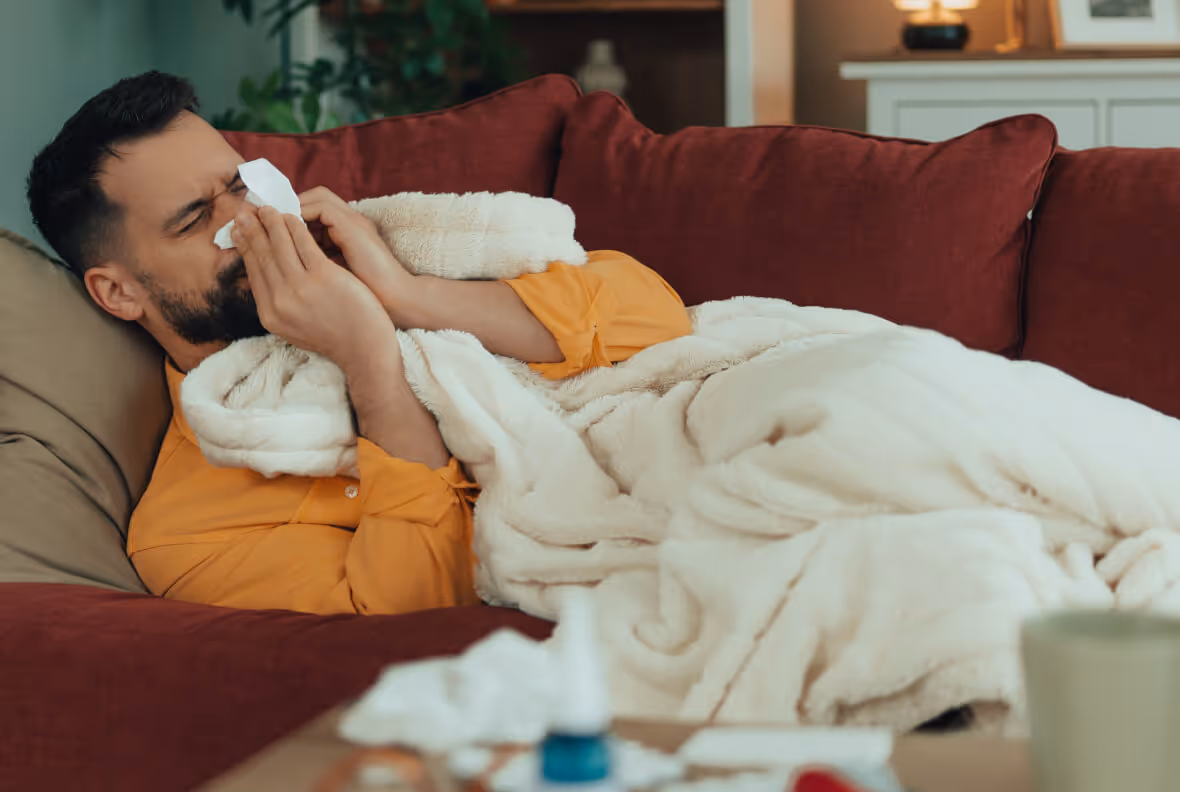 Man in an orange shirt lying on a red couch under a white blanket, blowing his nose with a tissue.