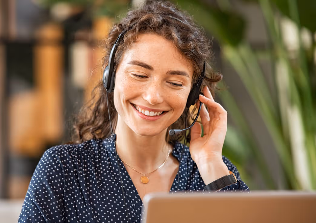 Smiling woman with curly hair wearing a headset and polka dot shirt looking at a laptop screen.