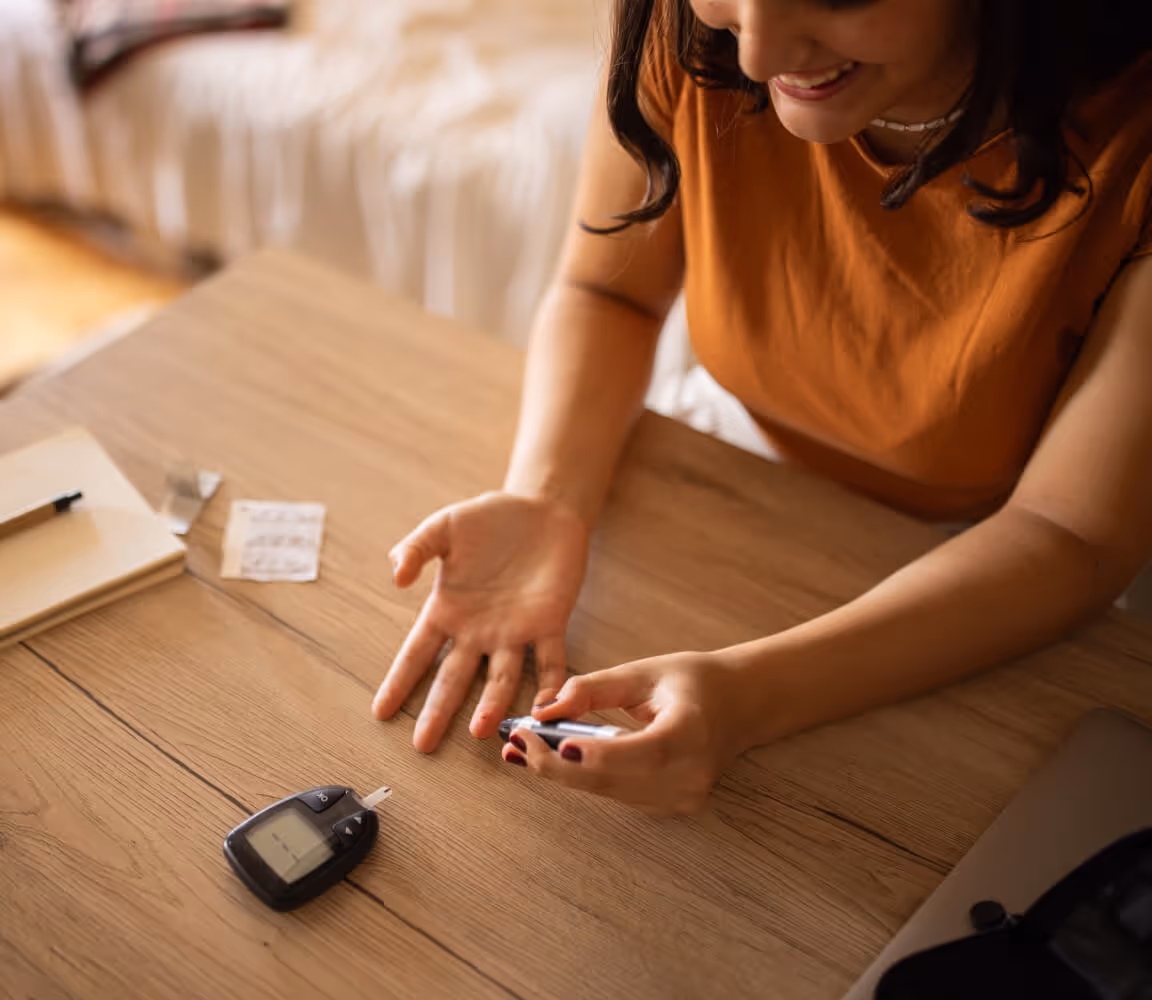 Smiling woman using a blood glucose meter to check her blood sugar at a wooden table.
