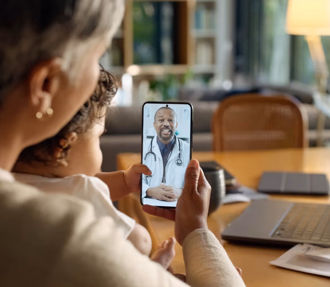 Adult holding a child while consulting a doctor on a smartphone during a video call at home.